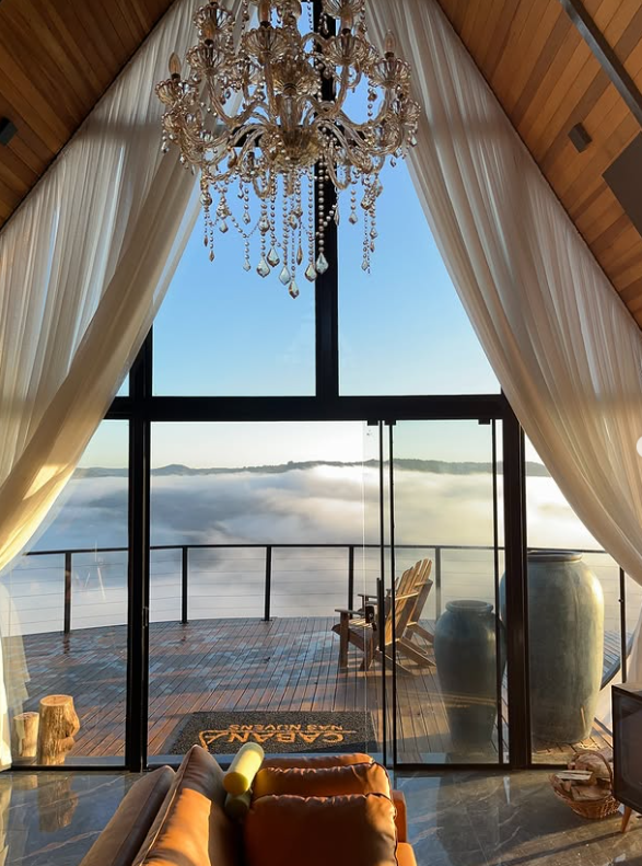 Vista interna da cabana com lustre, cortinas e deck suspenso acima das nuvens